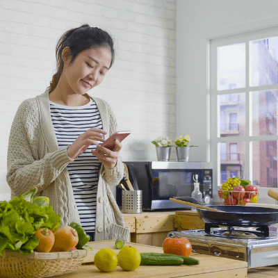 Young Asian woman cooking vegetables in wooden kitchen while looking at recipe on smartphone